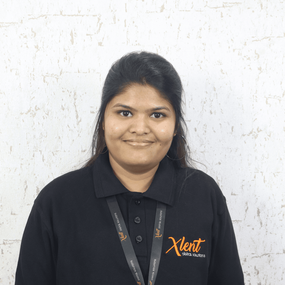 Jaysree Portrait of a smiling woman with dark hair, wearing a black polo shirt with an orange logo and a lanyard, standing against a light textured wall.