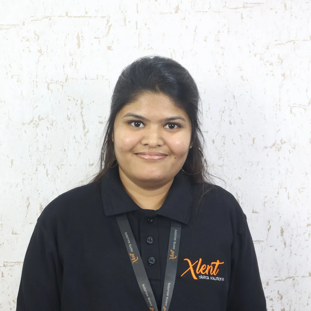 Portrait of a smiling woman with dark hair, wearing a black polo shirt with an orange logo and a lanyard, standing against a light textured wall.